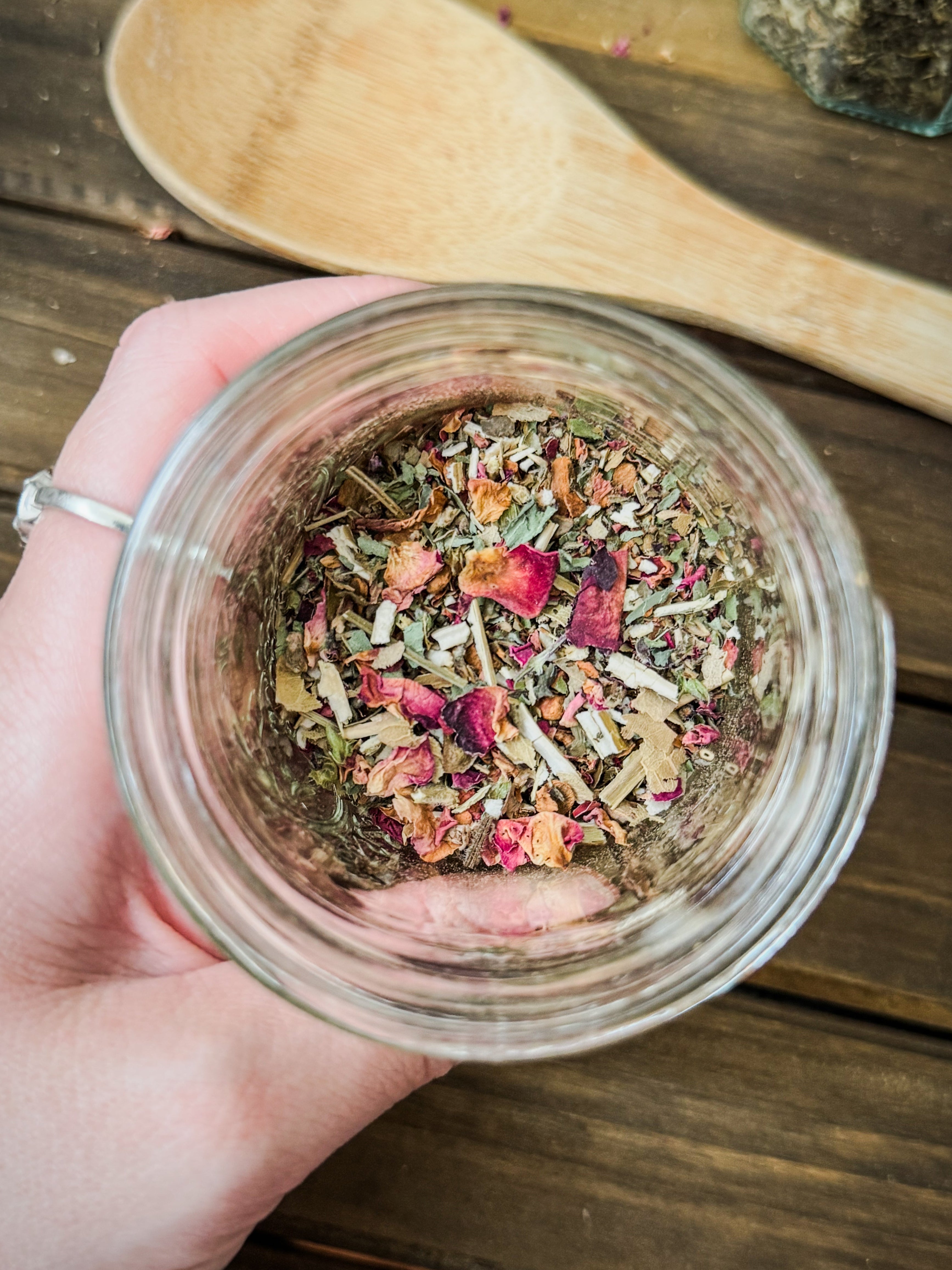 Hand holding a jar of dried herbs and flowers on a wooden surface with a wooden spoon.